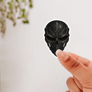 Pode incluir: Adesivo preto com um design detalhado de máscara de caveira. A máscara apresenta padrões intrincados e uma aparência escura e texturizada. O adesivo é segurado contra um fundo branco.
