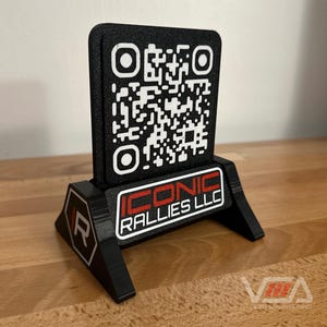 May include: A black 3D printed stand with a black and white QR code and the text "ICONIC RALLIES LLC" in red and white lettering.