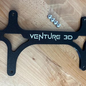 May include: Black, laser-cut metal bracket with the text "VENTURE 3D" in white. The bracket has a unique shape with multiple mounting holes and is accompanied by several small silver screws. The item is displayed on a wooden surface.