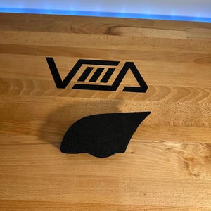 May include: A black textured grip pad with a curved shape, positioned below a black logo that reads "VMA" on a light wood surface. The logo is a stylized design with geometric shapes.