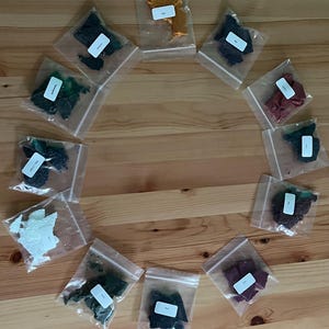 May include: A circular arrangement of colourful, irregularly shaped items, possibly wax melts or soap samples, each in a clear plastic bag with a white label. The colours include green, orange, red, and purple. The items are displayed on a wooden surface.
