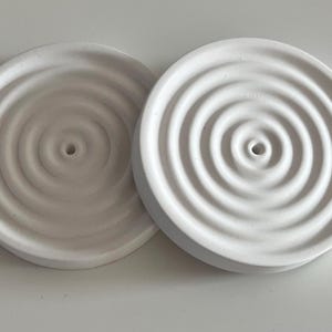 Op de afbeelding: Twee witte, cirkelvormige zeepbakjes met concentrische rimpelontwerpen. Elk bakje heeft een centraal afvoergat. Eén bakje is een lichtere tint wit dan het andere. De bakjes zijn gemaakt van keramiek of een vergelijkbaar materiaal.