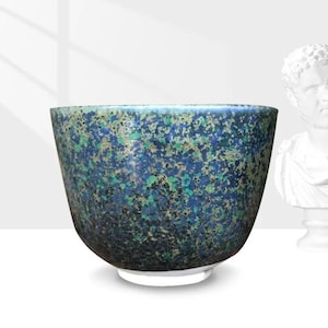 Op de afbeelding: Een gespikkelde keramische kom met een brede opening en een witte basis. De buitenkant van de kom heeft een gevlekt patroon van blauw, groen en zwart, waardoor een textuur ontstaat. De kom is een decoratief item.