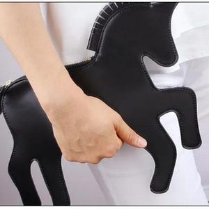 May include: A black clutch shaped like a unicorn silhouette with a smooth finish and a gold-toned zipper closure along the top edge. The bag features a distinct mane and tail detail, with stitched edges outlining the entire animal-shaped form.