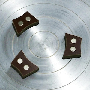 May include: Three brown, diamond-shaped magnets with two silver circular magnets each, arranged on a silver, circular surface. The magnets have a curved design and are evenly spaced.
