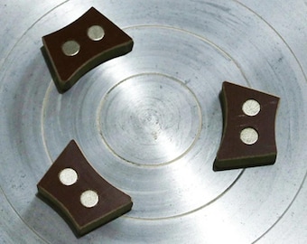 Può includere: Tre magneti marroni a forma di diamante, ciascuno con due magneti circolari argentati, disposti su una superficie circolare argentata. I magneti hanno un design curvo e sono uniformemente distanziati.