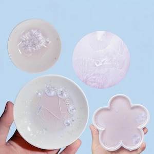 May include: A collection of small, light pink and white ceramic dishes and a flower-shaped tray. Each piece features delicate, crystal-like formations. The background is a soft, light blue, creating a serene and artistic presentation.