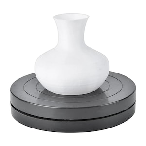 Puede incluir: Un jarrón de cerámica blanco con cuerpo redondeado y cuello esbelto, sobre un torno de alfarero negro giratorio. El jarrón tiene un acabado liso y mate. El torno es circular y tiene una superficie acanalada.