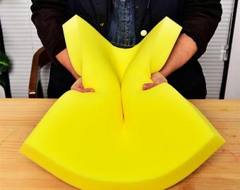 May include: A large, bright yellow foam cushion, held by a person. The cushion is shaped with rounded edges and a central indentation. The foam appears soft and flexible, with a smooth surface. The background is a wooden table.