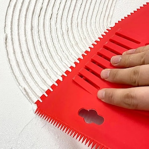 Könnte beinhalten: Ein rotes Kunststoff-Fliesennivellierwerkzeug mit einem gezackten Rand und einer Reihe von Rippen auf der Oberfläche. Das Werkzeug wird verwendet, um eine dünne Schicht weißen Klebstoffs auf einer Oberfläche zu verteilen.