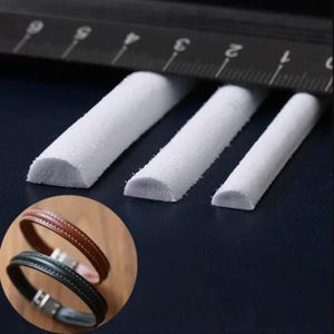 May include: Three white foam strips of varying lengths, each with a rounded edge. The strips are arranged in descending order of length, with the longest strip at the top of the image. The strips are on a black surface with a ruler visible in the background.