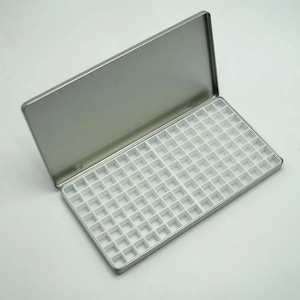 May include: An open, rectangular, silver-colored metal tin with a hinged lid. The interior features a white plastic grid with numerous small square compartments. The tin is designed for organizing small items, such as paints or beads.