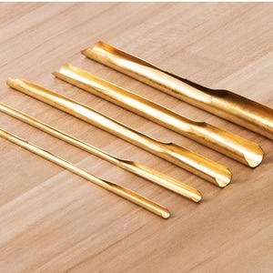 May include: Five gold metal tools of varying lengths, arranged in a row on a wooden surface. The tools are all hollow and have rounded ends.