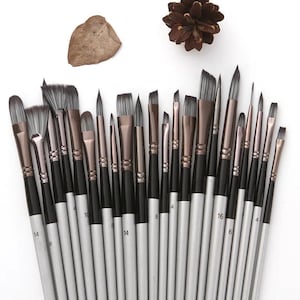 May include: A selection of artist paint brushes with black handles and silver ferrules. The bristles vary in shape and size, with some angled and others flat. The brushes are arranged on a white surface, with a pinecone and leaf in the background.