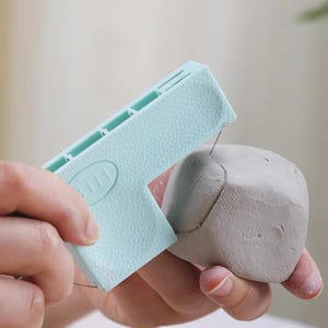 May include: A light blue clay cutter with a textured surface is used to slice a gray lump of clay. The cutter has a series of rectangular openings and the word "3111" is embossed on the side. The tool is held in the hand.