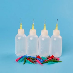 Puede incluir: Cuatro botellas exprimibles de plástico transparente con puntas amarillas y agujas negras, junto con una colección de puntas aplicadoras de colores. Las botellas tienen tapas blancas y marcas de medición. El fondo es azul claro.