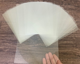 May include: A stack of clear plastic sheets, with one sheet being held by a hand. The sheets are translucent and appear to be made of a thin, flexible material.