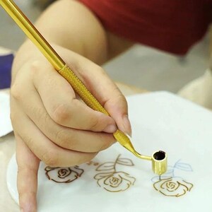 Puede incluir: Una herramienta dorada con forma de bolígrafo y mango texturizado se utiliza para dibujar diseños florales marrones sobre una superficie blanca. La herramienta tiene una punta curva con una pequeña abertura oscura, probablemente para tinta o pintura.