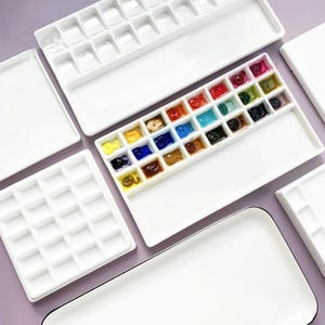 Puede incluir: Varias paletas de acuarela de cerámica blanca, algunas llenas de pintura colorida. Las paletas son rectangulares con pequeños pozos para mezclar colores. Suministros de arte.