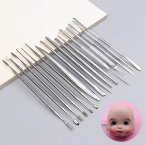 May include: A set of silver-coloured metal sculpting tools with various tips, including spatulas, points, and rounded ends, arranged in a diagonal row. A small, pale, round doll head with dark eyes and rosy cheeks is shown in the bottom right corner.