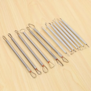 Pode incluir: Um conjunto de onze ferramentas de escultura com alças de arame metálico nas extremidades. Cinco ferramentas têm cabos de metal prateado com detalhes em laranja, enquanto seis ferramentas têm cabos cinza-claros. As alças variam em forma, incluindo designs redondos, triangulares e em gota para trabalhos detalhados em argila.