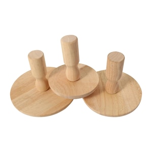 May include: Three wooden tampers with round bases and cylindrical handles. The tampers are made of light-colored wood and are arranged on a white background. The handles are attached to the center of the round bases.