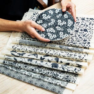 Op de afbeelding: Een ronde plaat met een zwart-wit bloemenpatroon wordt boven een stapel decoratief papier gehouden. Het papier heeft verschillende zwart-wit botanische ontwerpen, waaronder bloemen en vlinders.