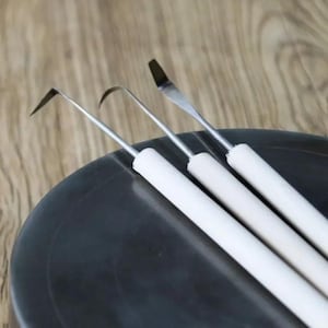 Puede incluir: Tres herramientas de cerámica de acero inoxidable con mangos blancos sobre una superficie gris oscuro. Las herramientas tienen puntas de diferentes formas: un ángulo puntiagudo, un gancho curvo y una espátula plana. Herramientas para esculpir y dar forma a la arcilla.