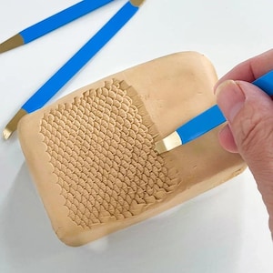 May include: A rectangular block of light tan clay featuring a textured pattern of small, overlapping scales carved into the surface. A blue-handled metal sculpting tool is being used to create the design, with another similar tool nearby.