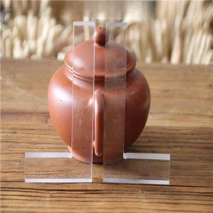 Può includere: Una teiera in terracotta con corpo arrotondato e manico sul lato. La teiera è esposta su due supporti in acrilico trasparente.
