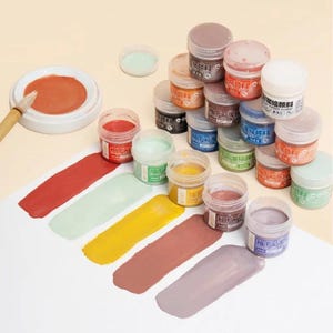 Puede incluir: Una colección de pequeños frascos de plástico redondos llenos de pigmentos de esmalte cerámico de varios colores. Varios frascos están apilados, mientras que otros están abiertos, mostrando su contenido. Cinco muestras de pintura son visibles.
