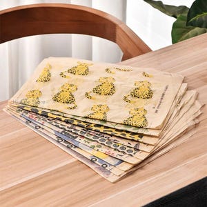 Può includere: Una pila di fogli di carta decorativi con vari motivi. I fogli superiori presentano un motivo ripetuto di illustrazioni di leopardi gialli su sfondo beige. Altri fogli mostrano disegni geometrici e astratti colorati. Le carte sono impilate su una superficie di legno.