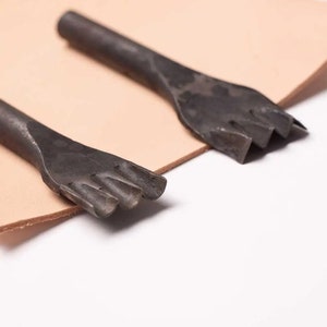 Pode incluir: Duas ferramentas de trabalho de couro de metal com lâminas de formas diferentes. As ferramentas estão sobre um pedaço de couro castanho claro.