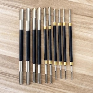 May include: A set of nine double-ended metal sculpting tools arranged in a row. Each tool features a black cylindrical handle, with some handles accented by gold-colored metal rings. The tips consist of various silver-toned shapes for carving.