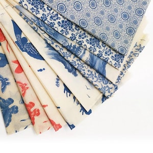 May include: A collection of folded fabric pieces with various blue and red patterns on a cream background. Designs include floral, bird, and landscape motifs. The fabrics are arranged in a fan shape, showcasing the different patterns.