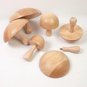 Puede incluir: Una colección de objetos de madera con forma de hongo en varios tamaños. Los hongos tienen sombreros y tallos de color marrón claro, con un acabado de veta de madera natural. Una pieza tiene el número "5.5" grabado. Los objetos están dispuestos sobre una superficie blanca.