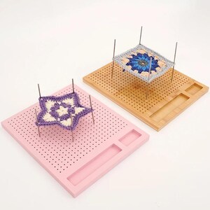 May include: Two square wooden blocking boards with grid holes and metal pins. One board is light pink with a purple and white star-shaped crochet motif. The other board is natural bamboo with a square blue, white, and grey crochet motif.