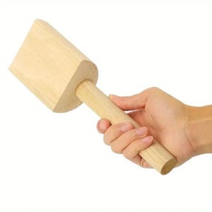 May include: A light-colored wooden mallet with a rectangular head and a cylindrical handle. The mallet is held in a hand against a white background. The tool appears to be new and unused.