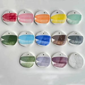 Könnte beinhalten: Eine Sammlung von fünfzehn weißen Keramikanhängern, jeweils mit einem einzigartigen abstrakten Pinselstrich-Design in verschiedenen Farben. Jeder Anhänger hat oben ein kleines Loch zum Aufhängen.