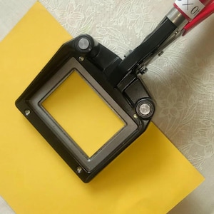 May include: A black and silver corner cutter tool with a red handle, positioned over a bright yellow paper. The tool has a rectangular opening and is designed for precise cuts. The background is a light-colored fabric.