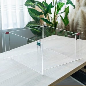 Puede incluir: Un marco de exhibición de acrílico transparente construido con cuatro paneles unidos en las esquinas con pequeñas pinzas metálicas. La estructura cuenta con dos pinzas rojas en los bordes exteriores y una pinza de color cerceta en el punto central.