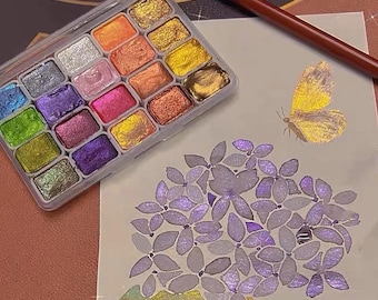 Puede incluir: Un juego de pintura de acuarela con una variedad de colores metálicos en una caja de plástico transparente. Un dibujo de una mariposa y una flor están en papel blanco, junto a un pincel marrón.