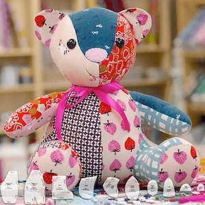 Könnte beinhalten: Ein Patchwork-Teddybär mit einer rosa Schleife, aus buntem Stoff mit Erdbeer- und Blumenmustern. Der Bär sitzt auf einem Tisch mit einem Satz Nähmustern.