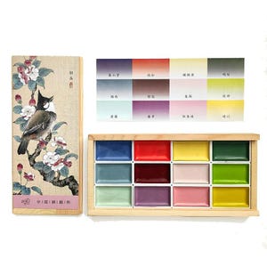 May include: A watercolor paint set with a wooden box featuring a bird and floral design. The set includes a palette of vibrant colors and a color chart with Chinese characters. The paints are arranged in a wooden tray.