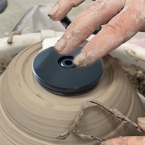 May include: A pottery wheel with a black centering disc on top. A hand is pressing down on the disc.