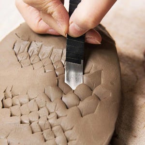 Op de afbeelding: Een hand die een klein metalen gereedschap gebruikt om een patroon in een stuk bruin klei te graveren.