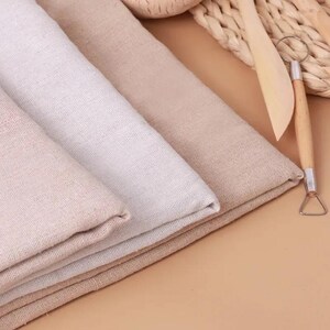 May include: Three folded pieces of woven fabric in varying shades of beige and off-white. A wooden pottery tool with a metal wire loop end rests on the right side, next to a woven wicker texture visible in the upper corner of the composition.