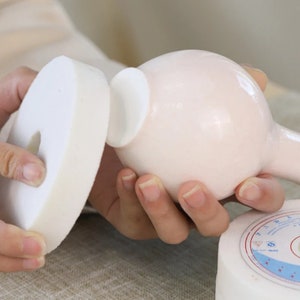 Puede incluir: Un jarrón de cerámica blanco con cuerpo redondo y cuello estrecho. El jarrón se está puliendo con una almohadilla de lijado blanca.