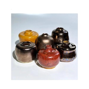 Puede incluir: Colección de seis jarrones de cerámica de varias formas y colores. Los jarrones van desde un bronce profundo hasta un rojo-naranja vibrante, con un acabado brillante. Algunos tienen una boca ancha, otros son más estrechos.