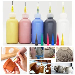 Puede incluir: Cinco botellas de pintura con puntas de aguja adjuntas, en amarillo, rosa, azul, gris y blanco. Las botellas están dispuestas en una fila de izquierda a derecha. Debajo de las botellas hay cinco puntas de aguja en negro, verde, rojo, azul y amarillo. La imagen también muestra varios ejemplos de cerámica decorada con la pintura.